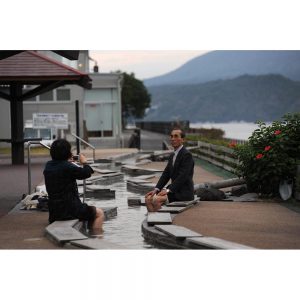 A couple takes pictures with Sakurajima behind while they put their feet in Ashiyu. Kagoshima, Kyushu, Japan
